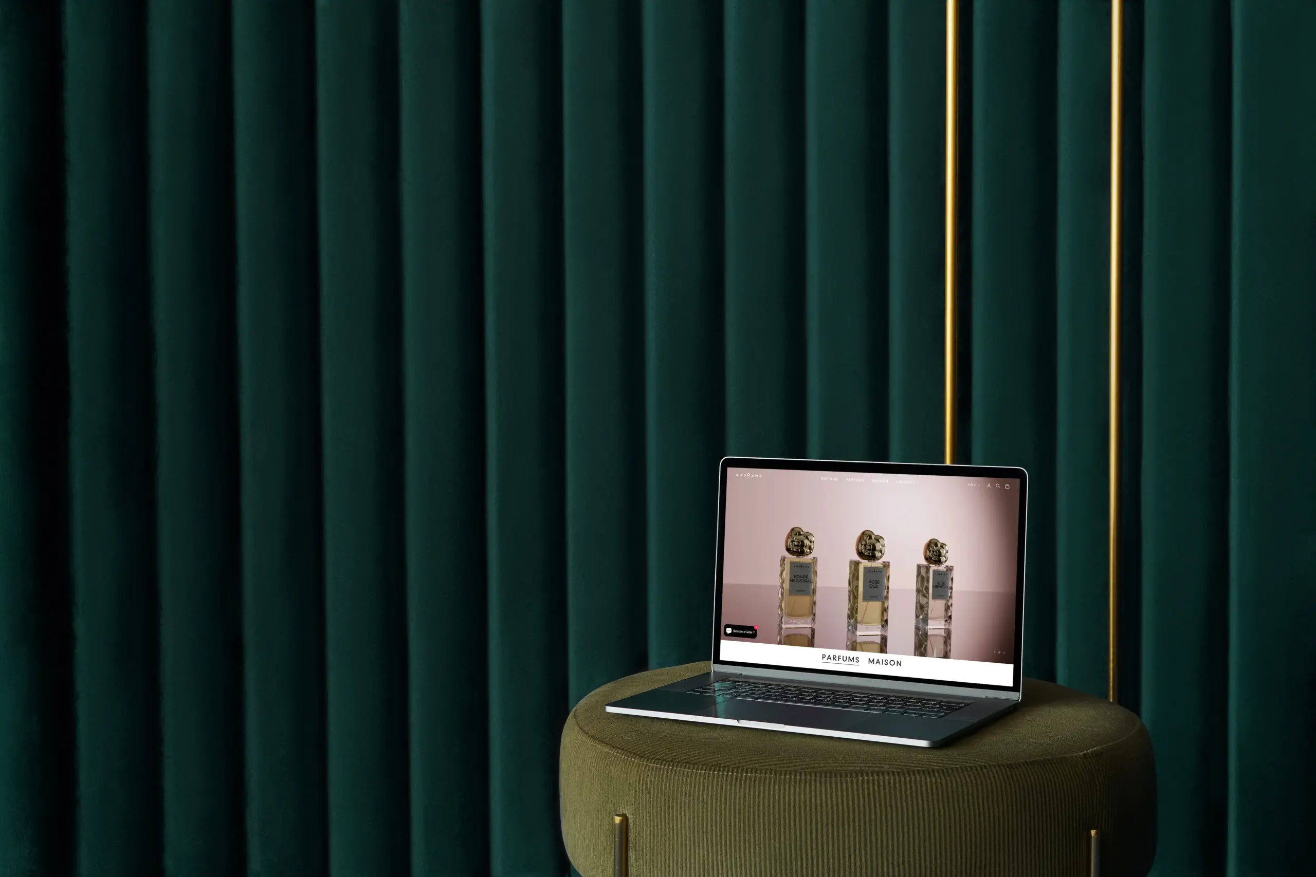 Mockup d’un site de parfumerie haut de gamme affiché sur un ordinateur portable posé sur un pouf vert, devant un rideau en velours vert foncé – ambiance élégante et minimaliste.