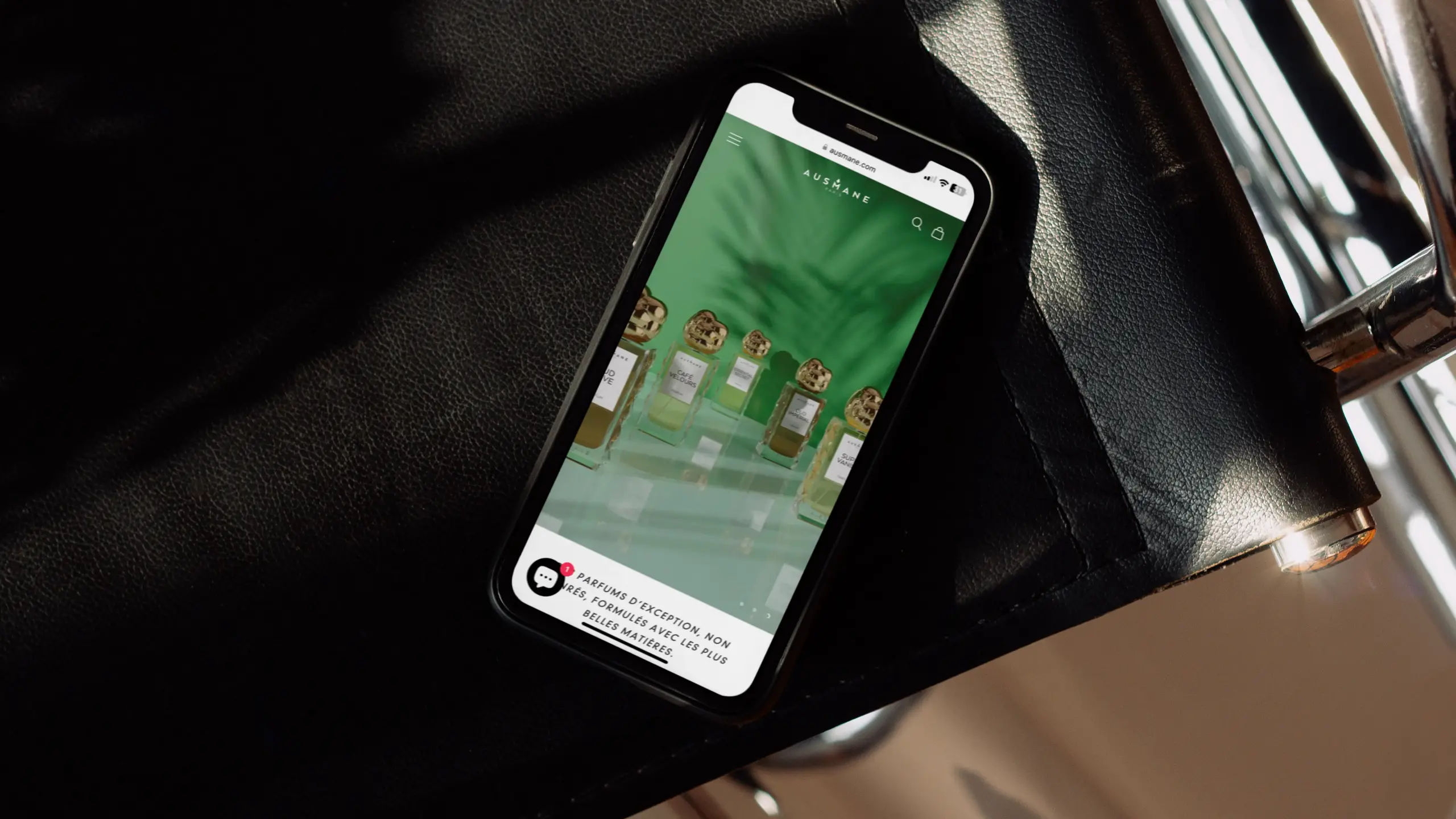 Affichage mobile du site ausmane.com sur un iPhone posé sur une chaise noire, mettant en avant une collection de parfums de niche avec arrière-plan vert végétal – design élégant et immersif.